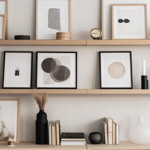 Puede incluir: Dos estantes de madera con obras de arte enmarcadas, velas, jarrones y libros. Las obras de arte incluyen diseños abstractos y un retrato en blanco y negro. Los estantes están contra una pared blanca.
