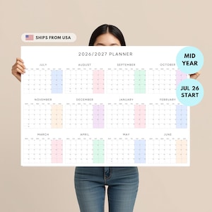 May include: A large white 2026/2027 planner with a colorful monthly layout. The planner includes the text "Mid Year" and "Jul 26 Start". The planner is held by a person.