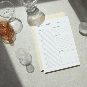 The daily schedule planner printed on Letter size paper with fine grid lines and clean typography, photographed on a neutral beige background with a coffee cup in the corner.