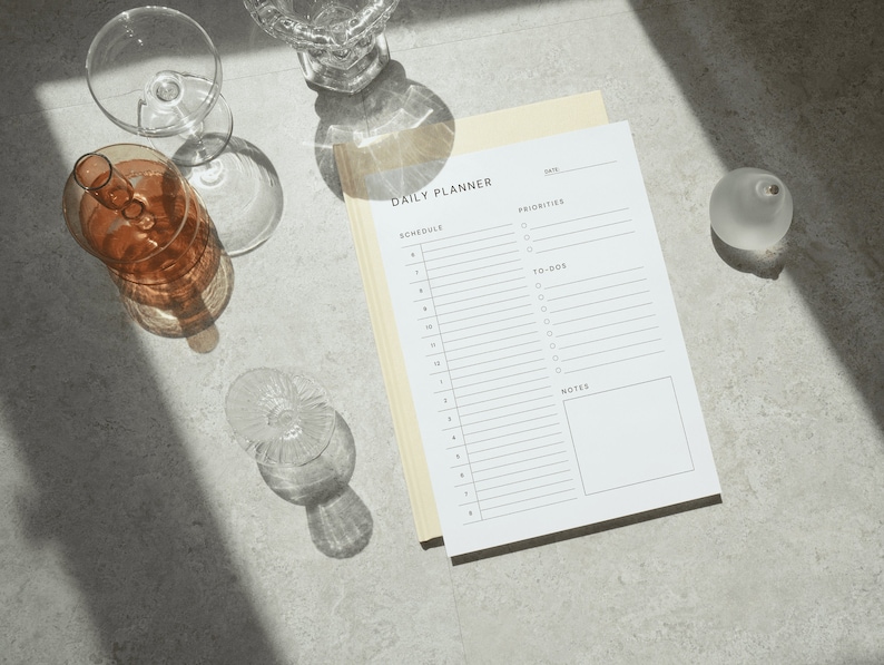 The daily schedule planner printed on Letter size paper with fine grid lines and clean typography, photographed on a neutral beige background with a coffee cup in the corner.
