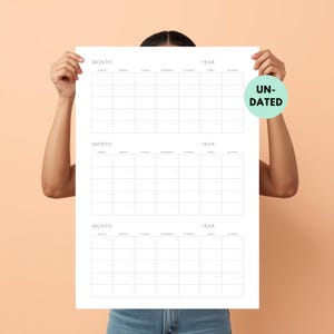Op de afbeelding: Een grote papieren wandkalender in wit met drie lege maandoverzichten met kopteksten voor maand en jaar. De kalender heeft dunne zwarte lijnen die het raster vormen en een ronde lichtgroene sticker met de tekst UN-DATED.