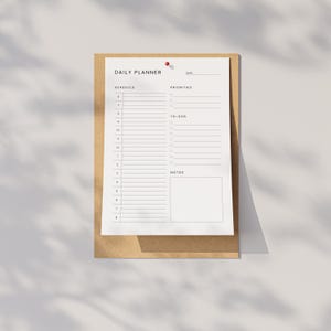 A printed PDF daily planner sheet on a flat white surface, showing date line, priorities, to-do list, and notes sections, next to a gold pen and paper clips arranged neatly.