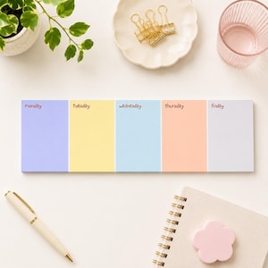 May include: A weekly planner with sections for each weekday, labelled Monday to Friday. The planner features a light blue, yellow, light blue, peach, and white colour scheme. A gold pen, a notebook, and a pink flower-shaped magnet are also visible.