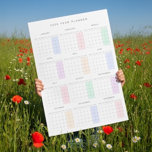 May include: A large 2026 year planner held in a field of wildflowers. The calendar is white with monthly layouts, each with a different pastel color highlight. The text "2026 YEAR PLANNER" is at the top.