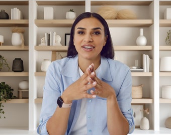 May include: A woman wearing a light blue button-down shirt is sitting in front of a white bookshelf with multiple shelves. The shelves are filled with various decorative items, including plants, vases, and books.