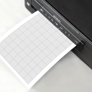 A white A4 sheet of printable 10x10 graph paper with fine grey grid lines, placed on a neutral desk background, showing the uniform ten-per-inch square pattern for drawing, writing, or technical sketches.