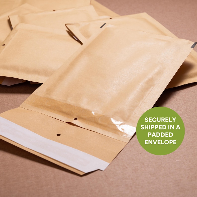 Puede incluir: Un mont&oacute;n de sobres acolchados marrones con una ventana transparente y una tira adhesiva blanca. El texto "SECURELY SHIPPED IN A PADDED ENVELOPE" est&aacute; en un c&iacute;rculo verde.