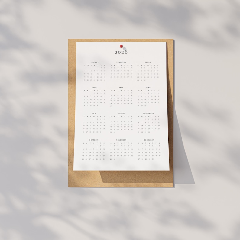 2026 printable calendar featuring all months in one page layout with a Monday start, printed in grey tones on A3 paper and taped to a white wall with a hand slightly adjusting the corner.