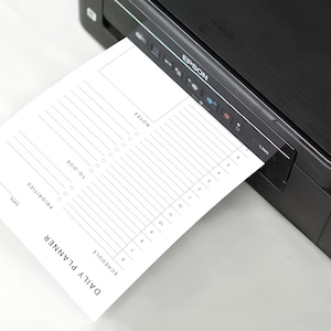 The minimalist planner page printed on smooth white paper, resting on a wooden desk beside a silver pen and phone, showing half-hourly schedule and priorities section.