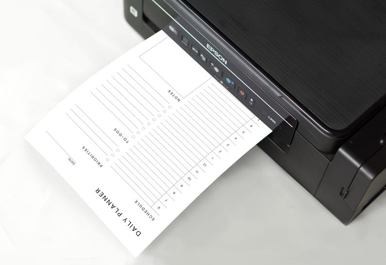 The minimalist planner page printed on smooth white paper, resting on a wooden desk beside a silver pen and phone, showing half-hourly schedule and priorities section.