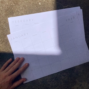 Minimalist 2026 calendar page, landscape orientation, grey lines and sans-serif font on white paper, placed on a neutral beige desktop.