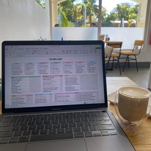 May include: A MacBook Air laptop displaying a spreadsheet titled "TO-DO LIST" sits open on a wooden table. A double-walled glass mug filled with a latte is next to the laptop. The background shows a window with a view of trees.