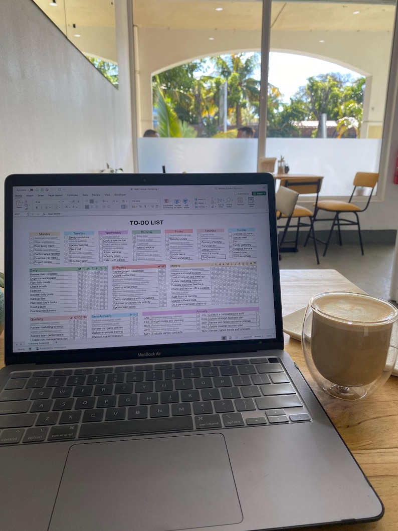 May include: A MacBook Air laptop displaying a spreadsheet titled "TO-DO LIST" sits open on a wooden table. A double-walled glass mug filled with a latte is next to the laptop. The background shows a window with a view of trees.