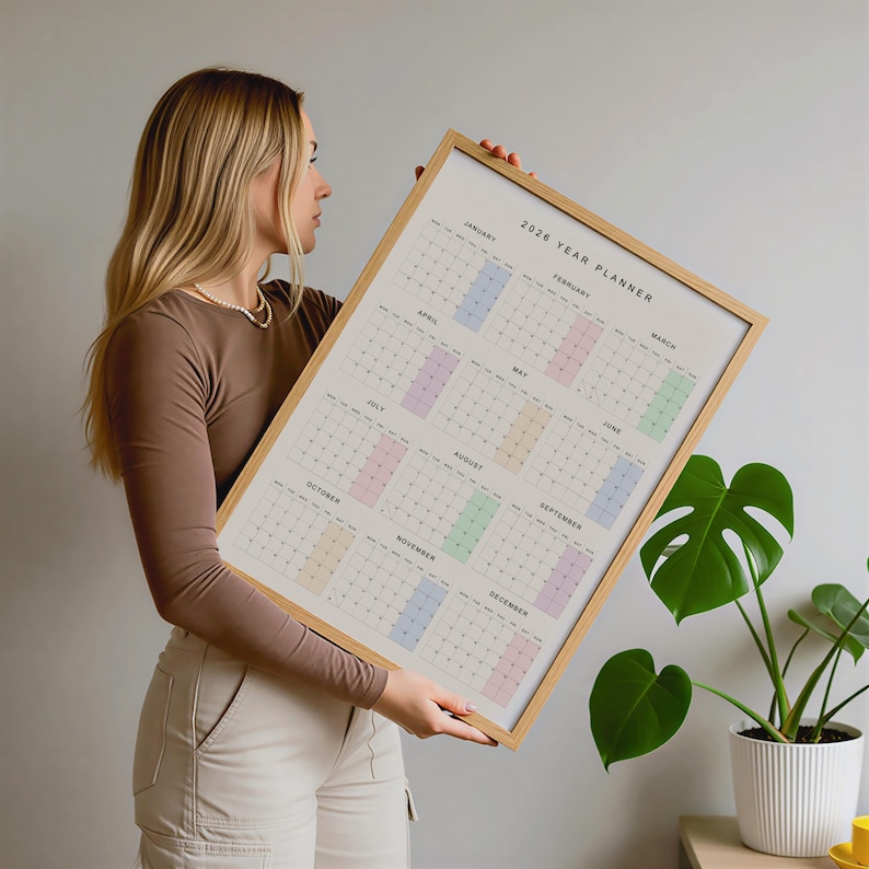 May include: A framed 2026 year planner with a light wood frame. The calendar grid features pastel color blocks for each month. The text "2026 YEAR PLANNER" is at the top. The calendar is held by a person.