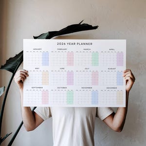 Op de afbeelding: Een 2026 jaarplanner wordt getoond. De kalender heeft een witte achtergrond met de vakjes van elke maand in een andere pastelkleur. De tekst "2026 YEAR PLANNER" staat bovenaan.