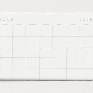 Close-up of a single 2026 monthly planner page in landscape format, grey modern typography and subtle lines on white paper held by a hand against a neutral wall.