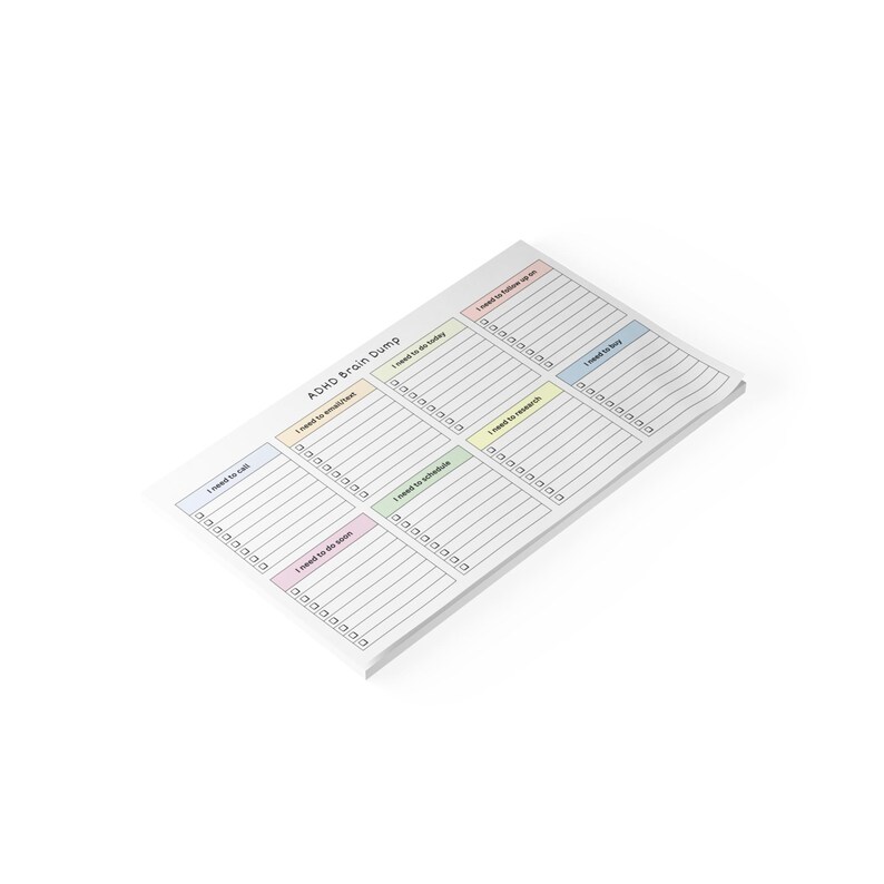 Top view of a 10x6 inch printed ADHD brain dump notepad with pastel colored section headings and checkboxes, placed on a desk with a pen, showing a clean productivity layout.