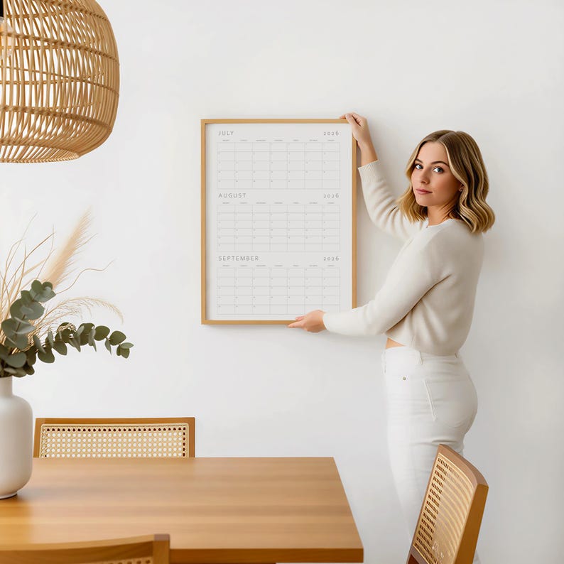 Op de afbeelding: Een ingelijste wandkalender met de maanden juli, augustus en september 2020. De kalender zit in een licht houten frame en wordt tegen een witte muur gehouden. De kalender heeft een minimalistisch ontwerp.
