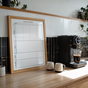 Puede incluir: Un planificador diario con marco de madera con el texto "DAILY PLANNER" se encuentra sobre una encimera de madera junto a una m&aacute;quina de espresso negra. Dos tazas con rayas verticales y un vaso transparente de caf&eacute; tambi&eacute;n est&aacute;n en la encimera. Plantas y azulejos blancos y negros en el fondo.
