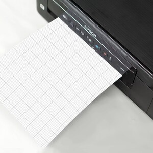 Stack of printable graph-paper sheets with 10 squares-per-inch grid, viewed from above, edges aligned neatly, showing repeated small square pattern across all pages.