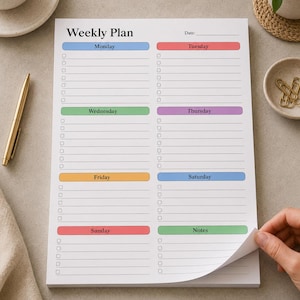 May include: A white weekly planner pad with sections for each day of the week, including Monday, Tuesday, Wednesday, Thursday, Friday, Saturday, and Sunday. Each day has a colored header and lined spaces for notes. The pad is on a neutral surface.