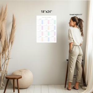 May include: A white 18"x24" 2024 year planner calendar with pastel-colored boxes for each month. The calendar is hanging on a white wall. A woman stands next to the calendar.