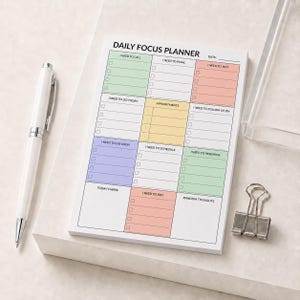 May include: A daily focus planner notepad with a white background and colorful sections for tasks, including headers like I Need To Call, I Need To Email, and Appointments. A white pen and a silver metal binder clip are placed next to the notepad.