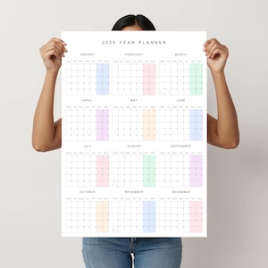 Pode incluir: Um grande planejador anual de 2026 com fundo branco. O calendário é dividido em meses, cada um com uma grade de datas. Os meses são codificados por cores com tons pastéis. O texto "2026 YEAR PLANNER" está no topo.