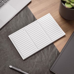 Può includere: Un blocco note bianco con linee e caselle di controllo grigie, appoggiato su una superficie grigio scuro. Sono visibili anche un laptop, una penna e una pianta grassa in un vaso scuro. Il blocco note è progettato per fare liste e organizzare.