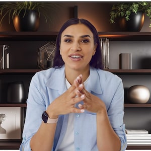 May include: A woman wearing a light blue button-down shirt sits in front of a bookshelf with dark wood shelves. The shelves are decorated with various objects, including plants, vases, and books.