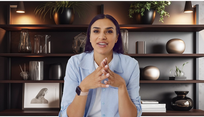 May include: A woman wearing a light blue button-down shirt sits in front of a bookshelf with dark wood shelves. The shelves are decorated with various objects, including plants, vases, and books.