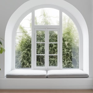 May include: A white window seat with a large arched window overlooking a green tree. The window seat is cushioned and has a white wall behind it. A potted plant is in the corner of the room.