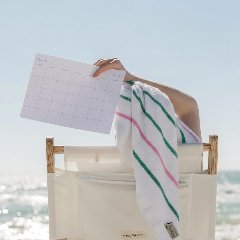 Person holding a printable 2027 monthly planner outdoors near the beach, showing the clean landscape calendar layout.