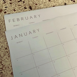 Flat lay of a printed 2026 calendar month on white A3 paper, landscape orientation with thin grey grid lines and clean sans-serif font for dates, placed on a light wooden table.