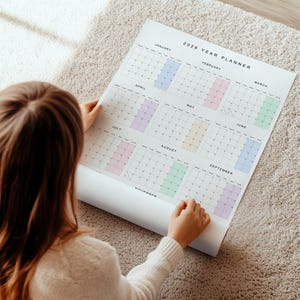 Overhead flat lay of 2026 wall calendar on white paper, portrait layout, vibrant pastel month headers, full-year grid visible, clean design showing weekdays and all dates clearly.