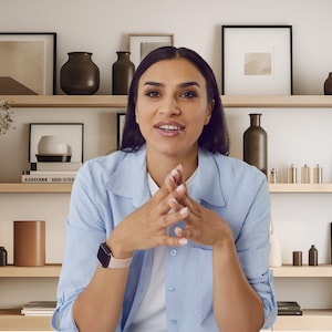Può includere: Una donna con una camicia abbottonata blu chiaro è seduta di fronte a una libreria con vari oggetti decorativi, tra cui vasi, opere d'arte incorniciate e libri.