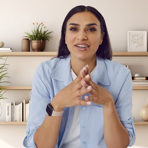 May include: A woman wearing a light blue button-down shirt with a watch on her left wrist. She is sitting in front of a white wall with wooden shelves behind her. The shelves are decorated with plants, books, and decorative objects.