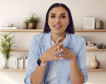 May include: A woman wearing a light blue button-down shirt with a watch on her left wrist. She is sitting in front of a white wall with wooden shelves behind her. The shelves are decorated with plants, books, and decorative objects.
