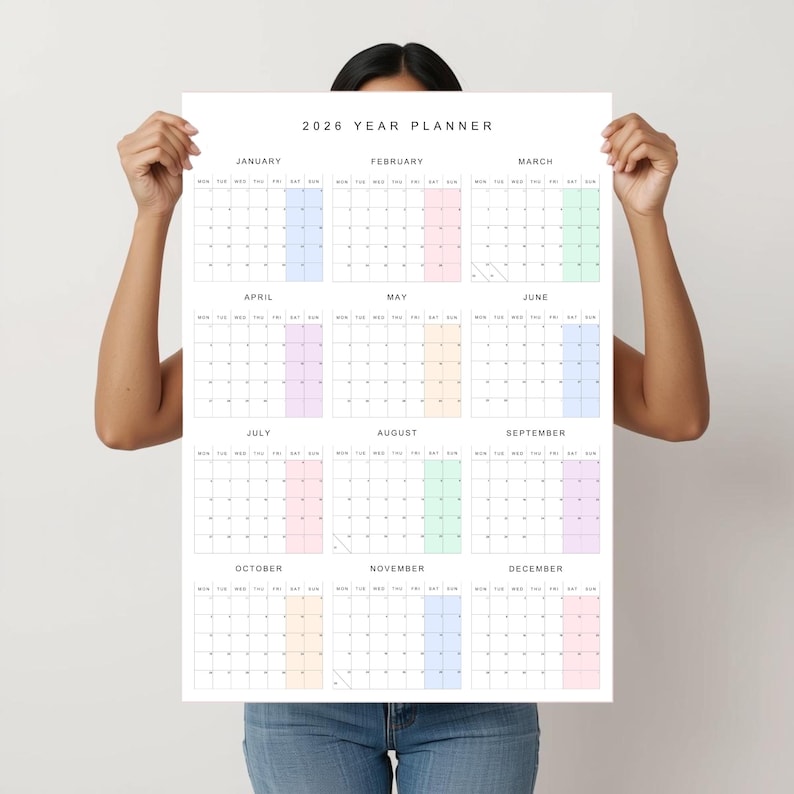 Vertical 2026 wall calendar shown taped to a neutral beige wall, printed on white paper with thin black lines, pastel-colored weekends, and a full year on a single page with Monday start.