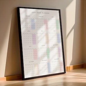 Vertical 2026 wall calendar shown taped to a neutral beige wall, printed on white paper with thin black lines, pastel-colored weekends, and a full year on a single page with Monday start.
