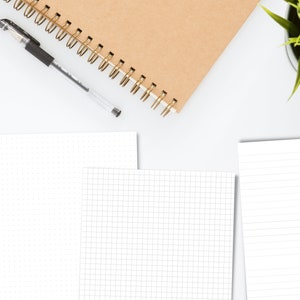 May include: A flat lay of a white desk with a brown notebook, a black pen, and three sheets of paper. The paper is blank and has different grid patterns. A potted plant is in the top right corner of the image.