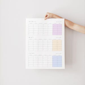 May include: A white calendar for April, May, and June 2026, with each month's days arranged in a grid. The 2026 dates are highlighted with pastel colors: purple, yellow, and blue. The calendar is held by a person's hand.