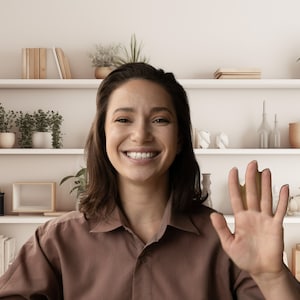Peut inclure: Une femme aux cheveux bruns sourit et salue de la main la caméra. Elle porte une chemise brune et se tient devant une bibliothèque blanche avec des plantes et des livres.