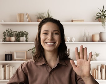 May include: A woman with brown hair smiles and waves at the camera. She is wearing a brown shirt and is standing in front of a white bookshelf with plants and books.
