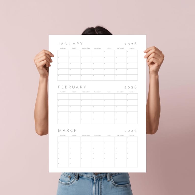 Op de afbeelding: Een witte papieren kalender voor het jaar 2026, met de maanden januari, februari en maart. De kalender wordt omhoog gehouden door een persoon die een blauw spijkeroverhemd draagt. De achtergrond is effen lichtroze.