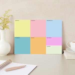 Op de afbeelding: Een kleurrijke weekplanner met pastelkleurige blokken voor elke dag van de week. De dagen van de week zijn in donkerblauwe letters beschreven. De planner is rechthoekig en heeft een witte achtergrond.