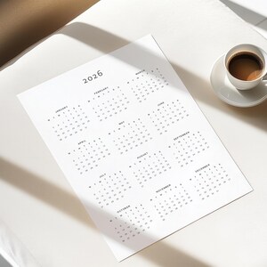 Year at a glance 2026 calendar printed on A4 paper with portrait orientation, featuring all months in a structured grid layout with minimalist grey font, styled on a wooden desk surface