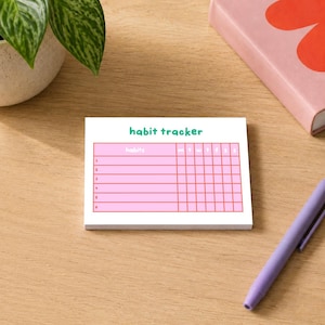 May include: A rectangular white paper notepad featuring a pink grid for tracking habits. The text "habit tracker" is printed in green at the top. The grid includes columns for days of the week labeled m, t, w, t, f, s, s and six numbered rows.