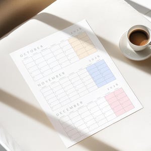 May include: A printed calendar for October, November, and December 2026, with color-coded boxes. A white ceramic cup and saucer with coffee sits to the right. The calendar is on a white surface.