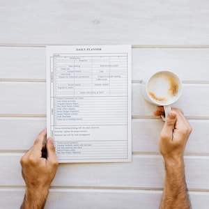 Puede incluir: Un planificador diario con un horario detallado y una lista de tareas es sostenido por una persona. Una taza blanca de caf&eacute; con una capa de espuma se sostiene en la otra mano. El planificador y la taza est&aacute;n sobre una superficie de madera blanca.